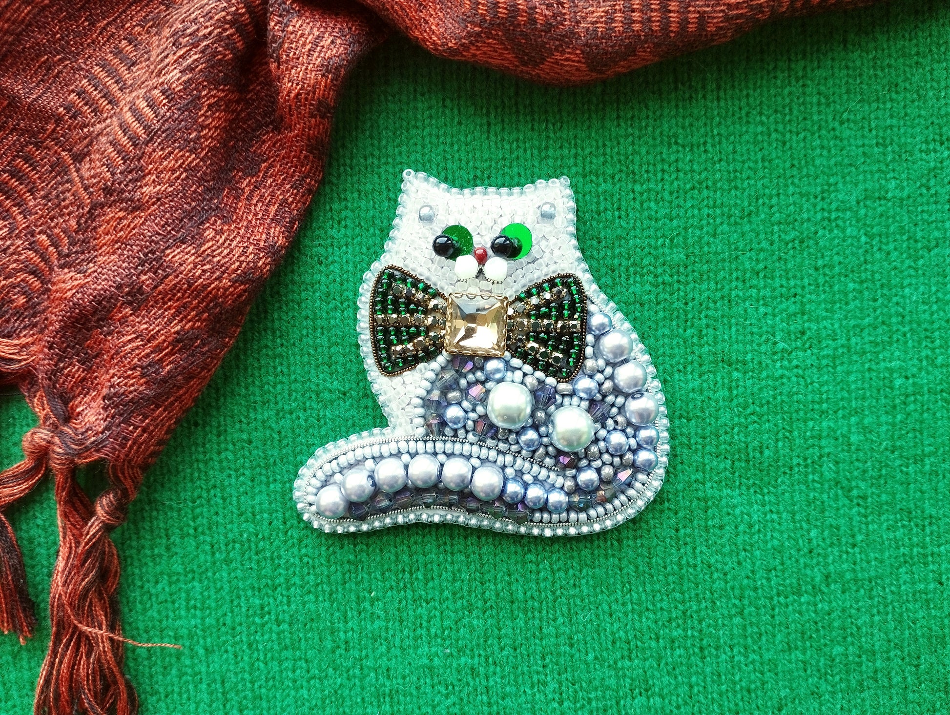 A decorative cat brooch made of beads and jewels, with a bow tie, is displayed on a green fabric surface.