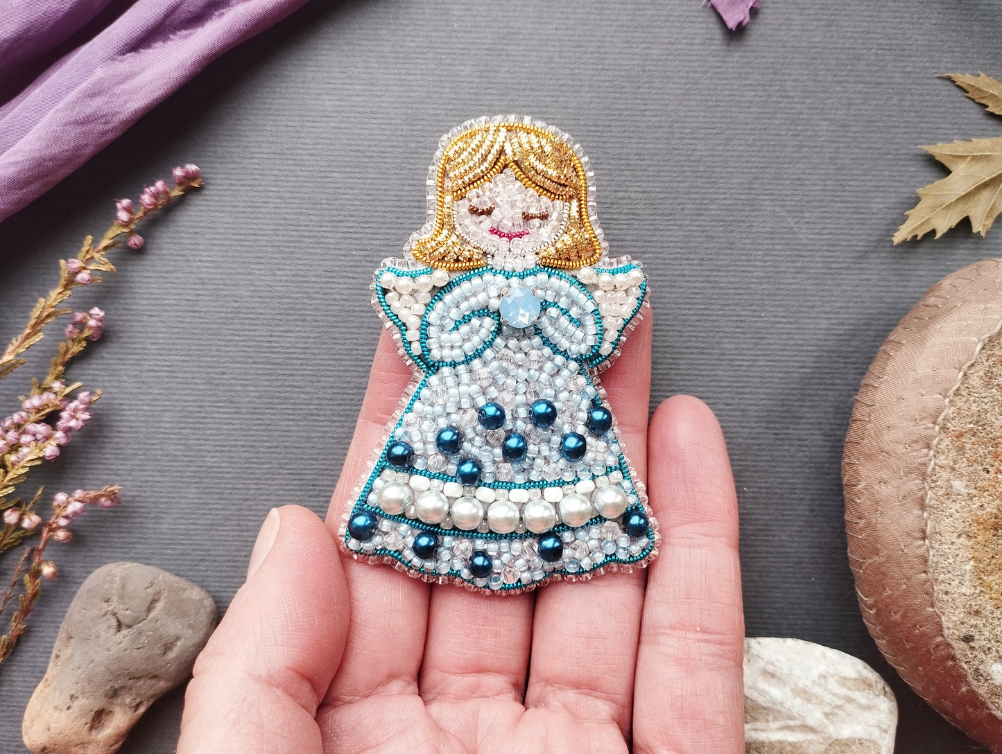 A hand holding a small, intricately beaded doll with a blue dress and white pearls, surrounded by various natural elements such as flowers, leaves, and rocks.