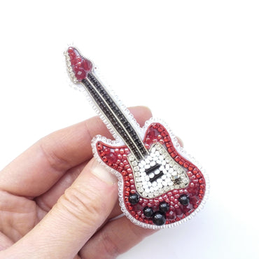 A hand holding a small, intricately designed embroidered guitar brooch.