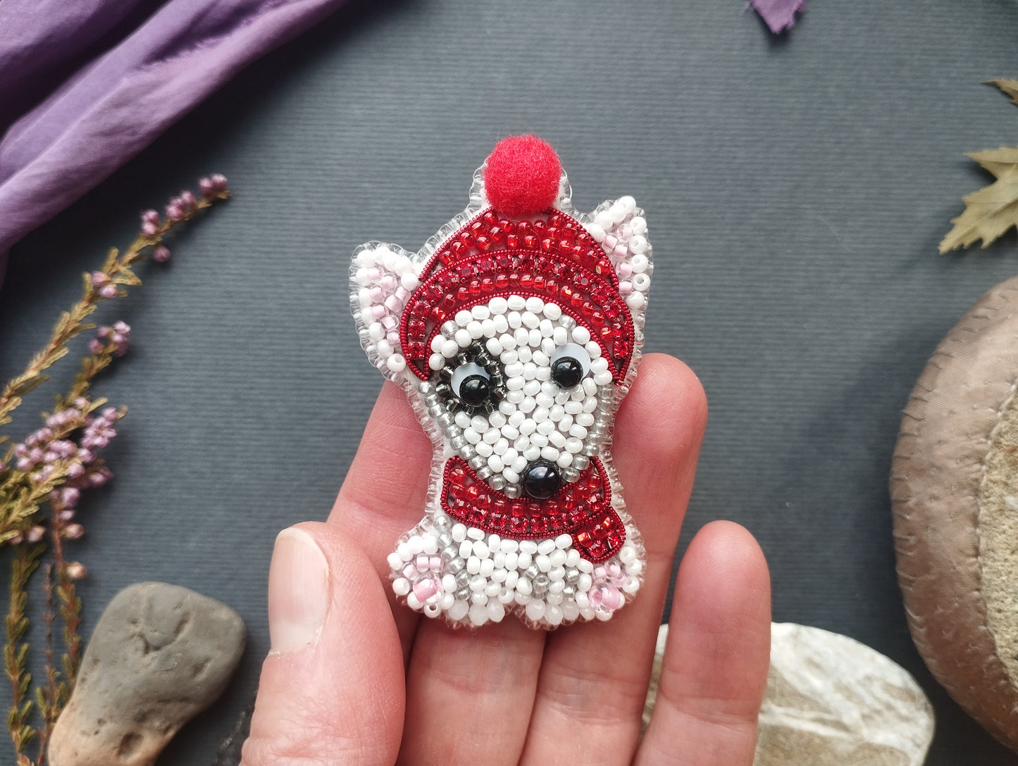 A hand holding a small, hand-made embroidered object resembling a dog wearing a red hat and scarf.