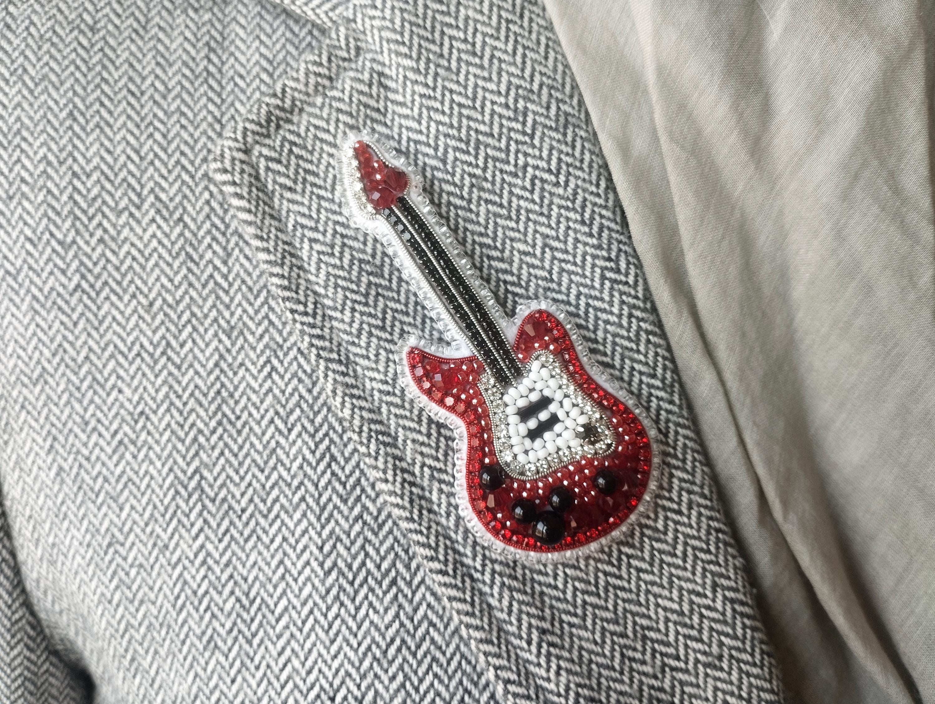 A close-up of a person wearing a grey tweed jacket with a red and white guitar-shaped brooch or pin attached to the lapel.