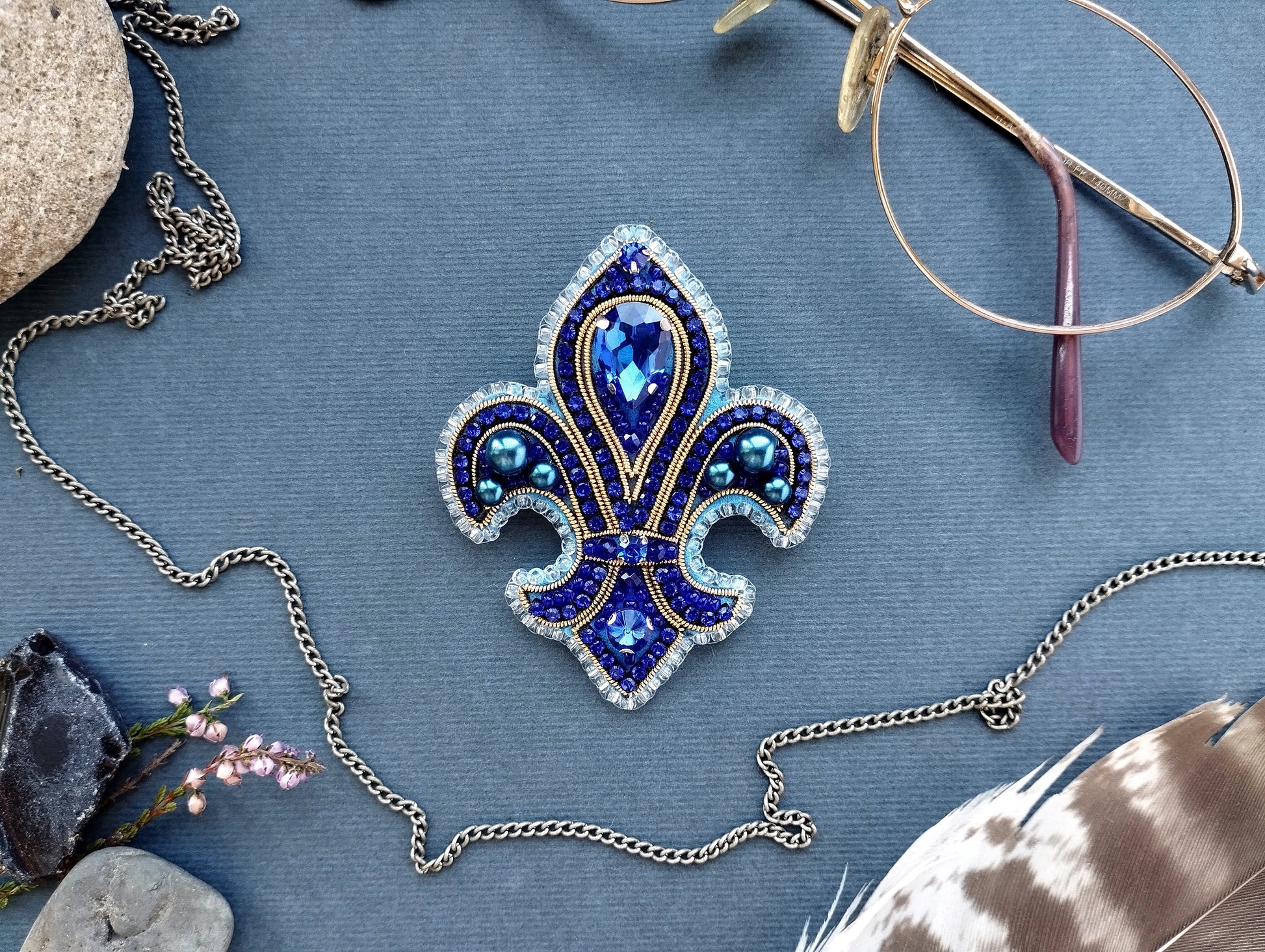 a blue and gold brooch with a blue and gold design, surrounded by various accessories such as a necklace, glasses, and feathers.