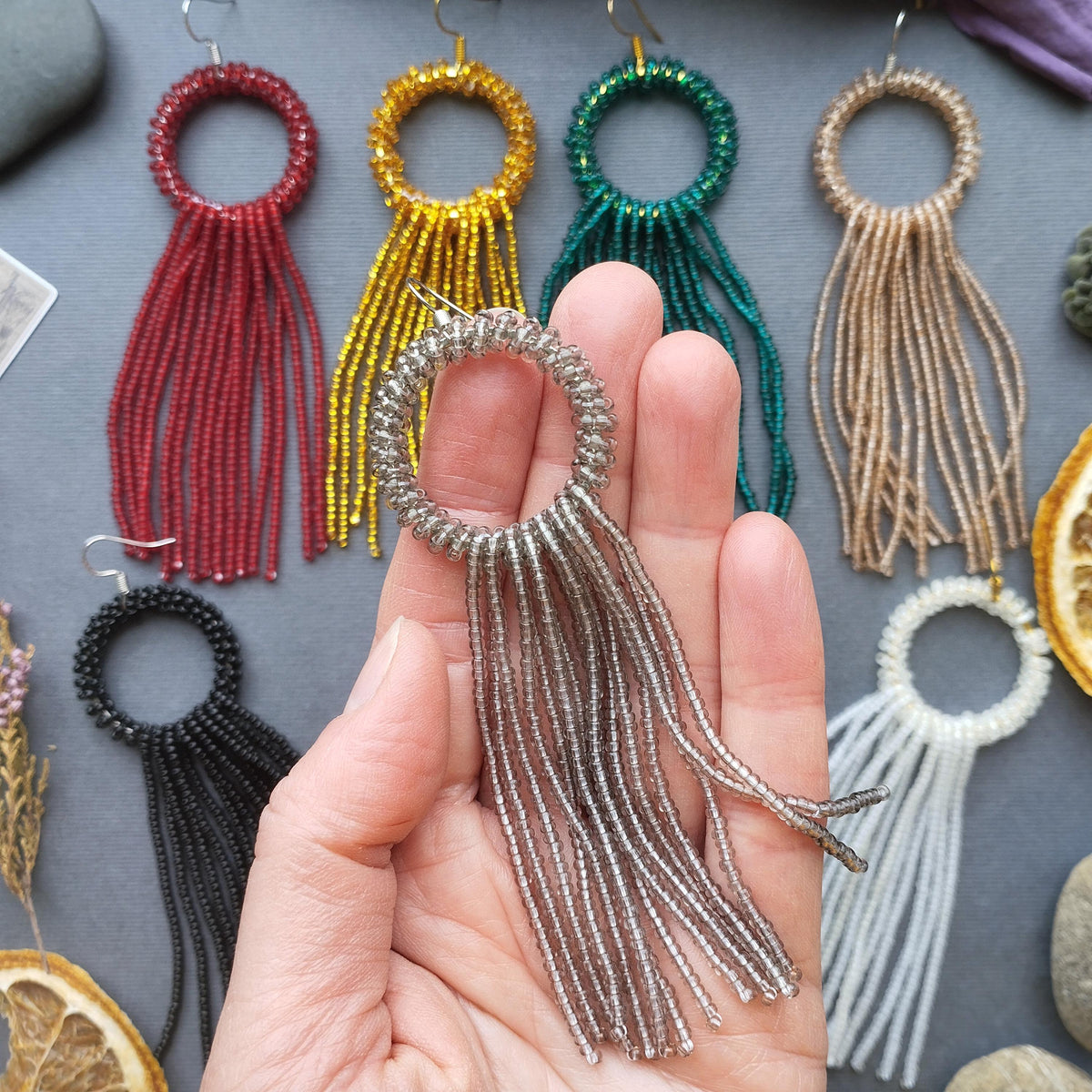 A hand holding a pair of silver earrings with long, dangling tassels in various colors, including red, yellow, green, and beige.