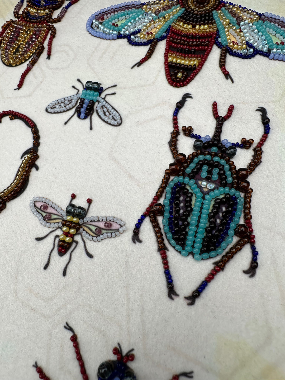a close-up view of several intricately crafted insects, including a large blue and green beetle, a smaller white and red bee, and a red and black ant. The insects are meticulously crafted using a variety of colorful beads and threads, showcasing the artist's skill and attention to detail.