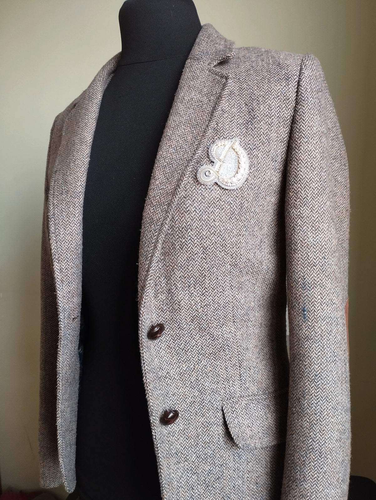 A gray tweed jacket with a white embroidered patch on the left chest area, displayed on a mannequin.