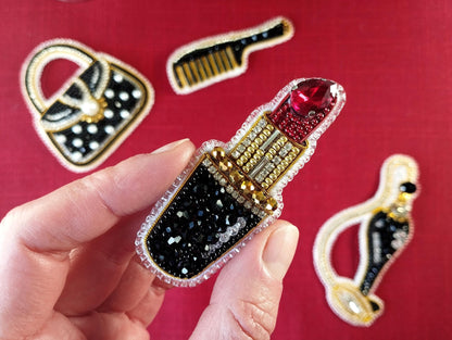 A hand holding a decorative pin featuring a red lipstick design, with various accessories such as a handbag, comb, and other embellishments visible in the background.