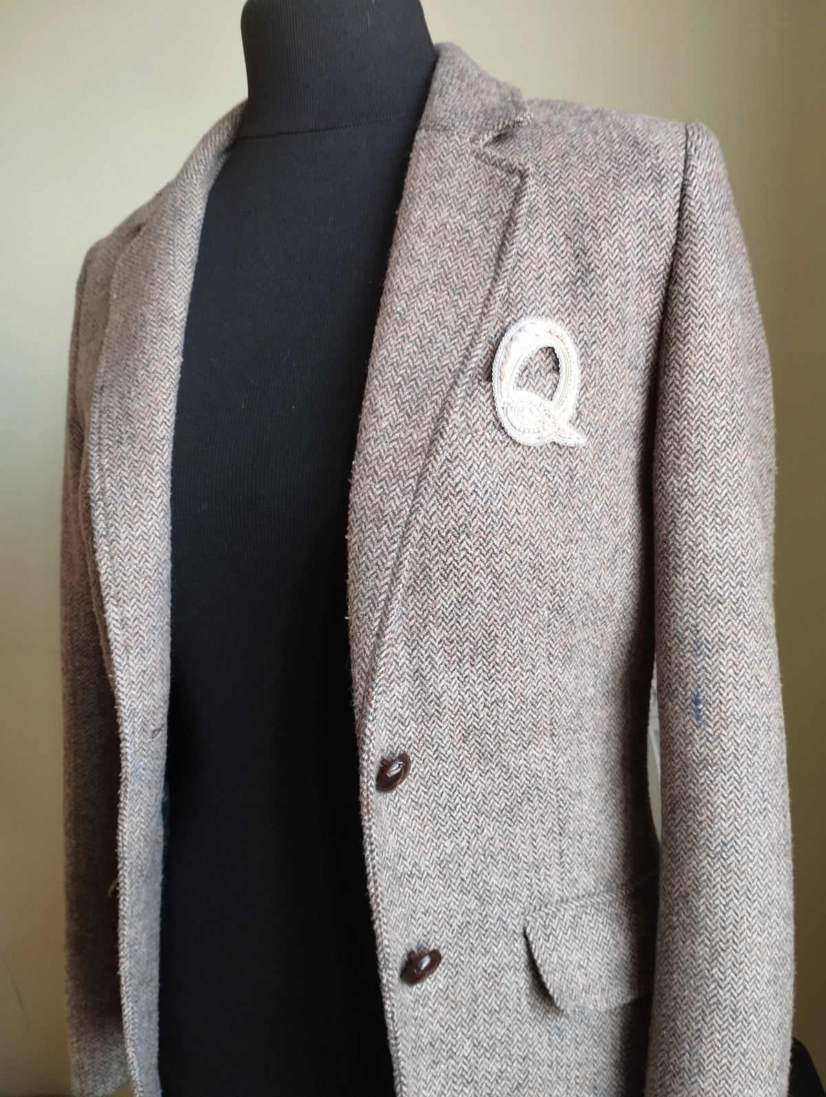 A gray tweed jacket with a white "Q" pin on the lapel.