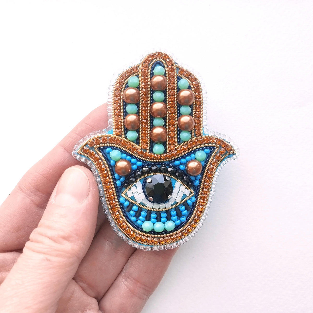 A hand holding a colorful, intricately designed button or brooch with a blue and gold color scheme, featuring a large eye-shaped embellishment.