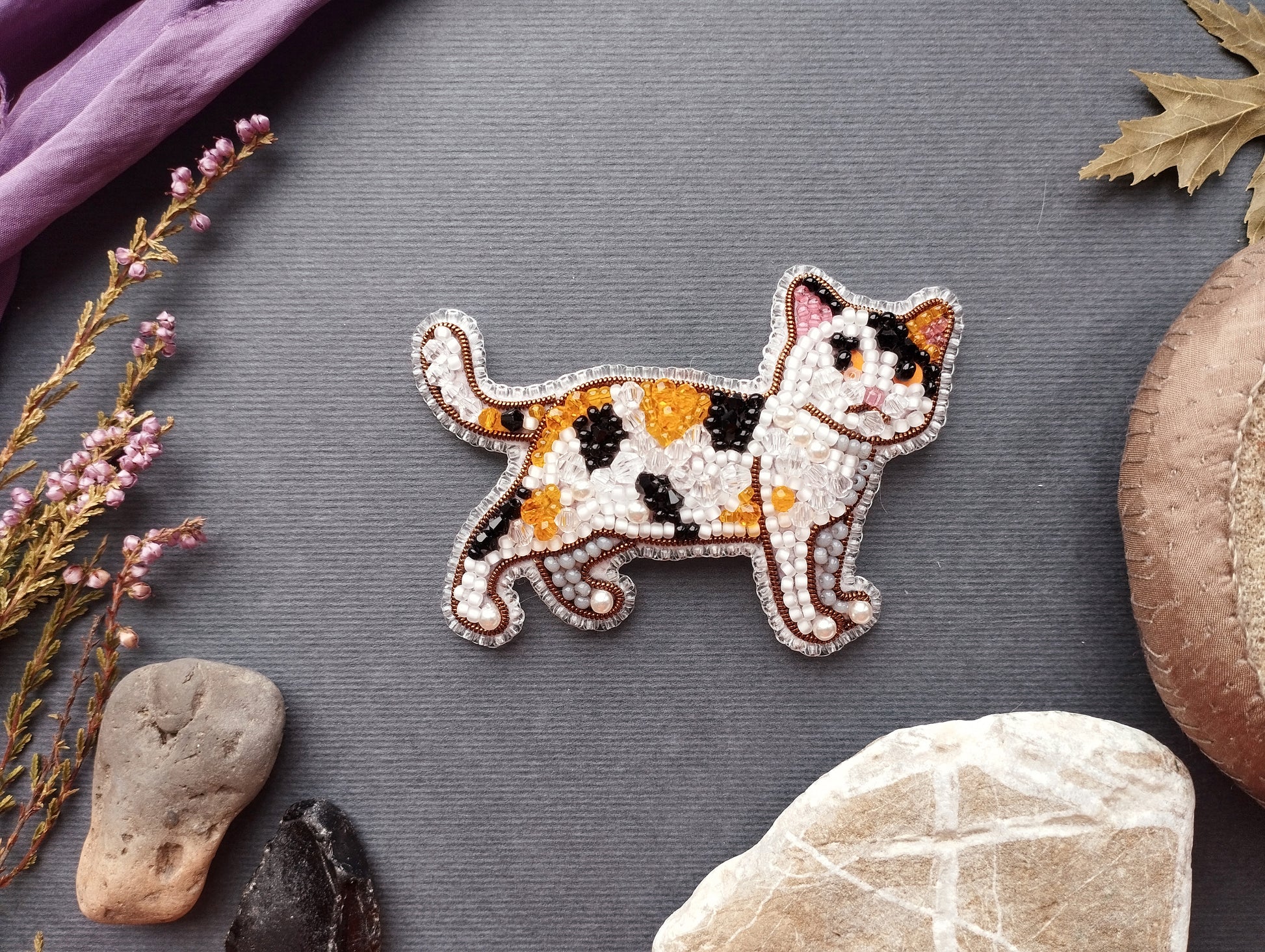 a decorative beaded cat brooch placed on a grey surface, surrounded by various natural elements such as rocks, leaves, and flowers.