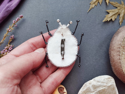 A hand holding a small, intricately designed insect-like object made of fabric, beads, and metal.