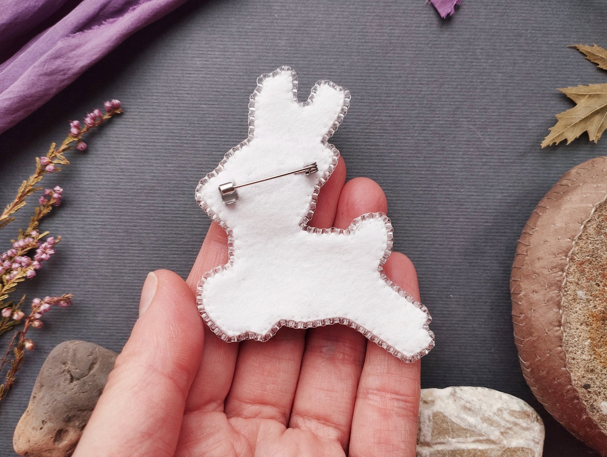 A hand holding a white felt rabbit pin against a dark background with various objects, including flowers and rocks.