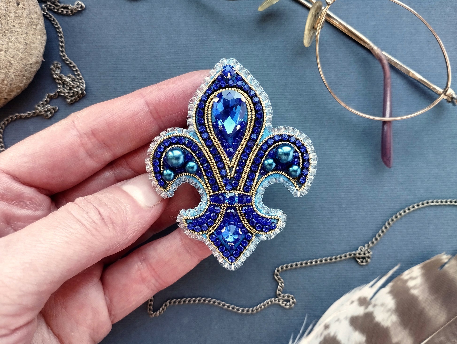 A hand holding a decorative blue and silver brooch with a large blue gemstone in the center, surrounded by smaller blue gemstones and intricate designs.