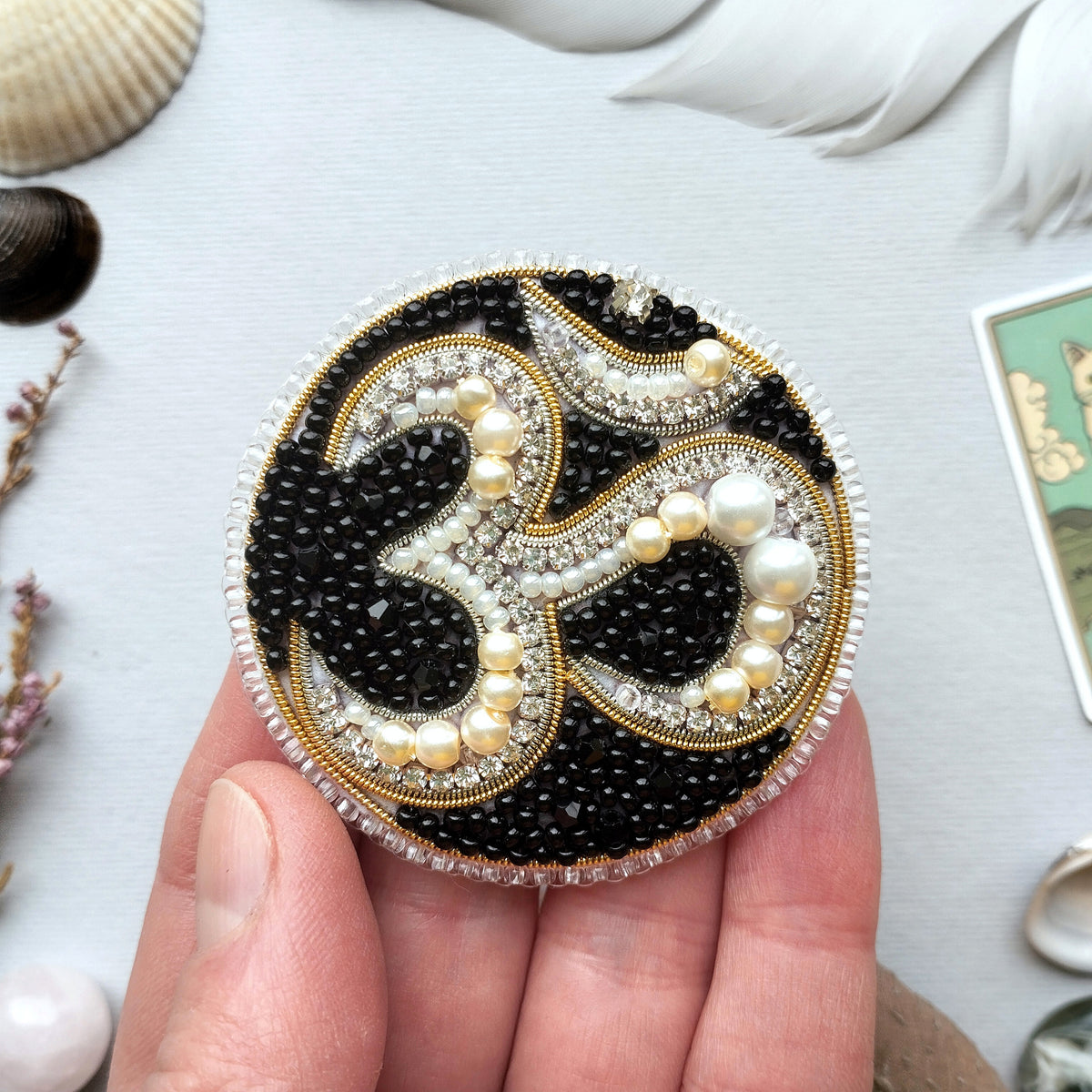 A hand holding a decorative piece with a black and gold design, featuring the Om symbol and pearls.
