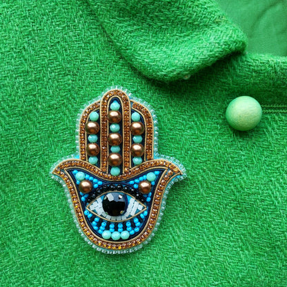 a close-up of a handcrafted brooch with a blue and gold design, adorned with a large eye and surrounded by small beads. The brooch is placed on a green fabric, which provides a contrasting background.