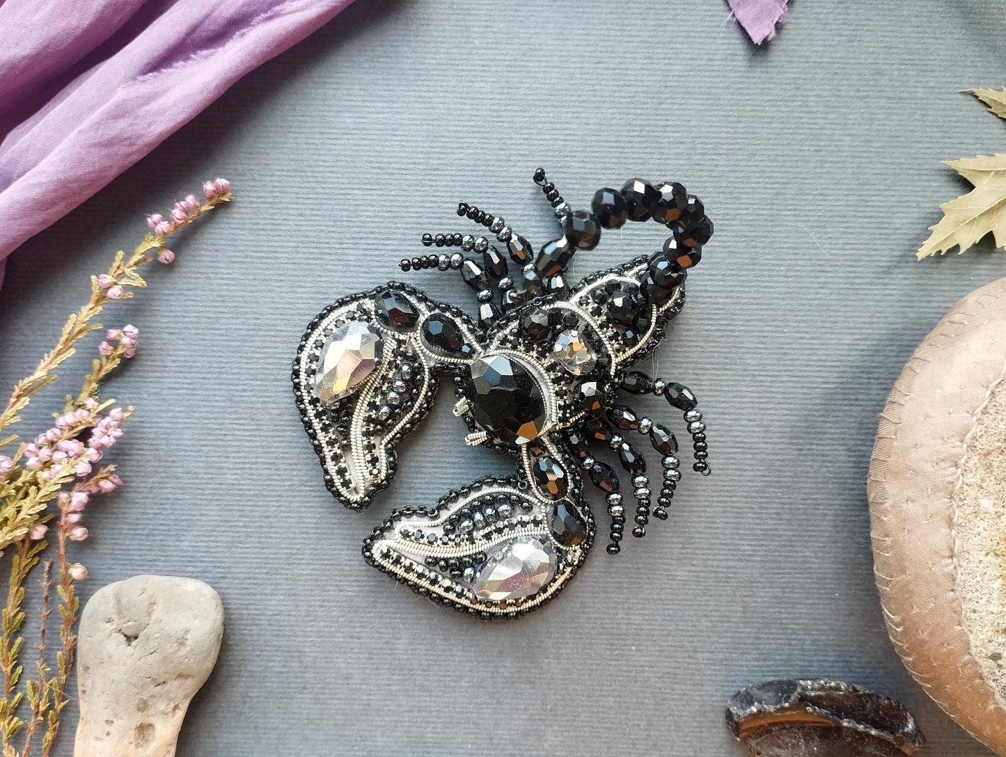 a black and silver brooch with a detailed design, surrounded by various natural elements such as flowers, leaves, and rocks.