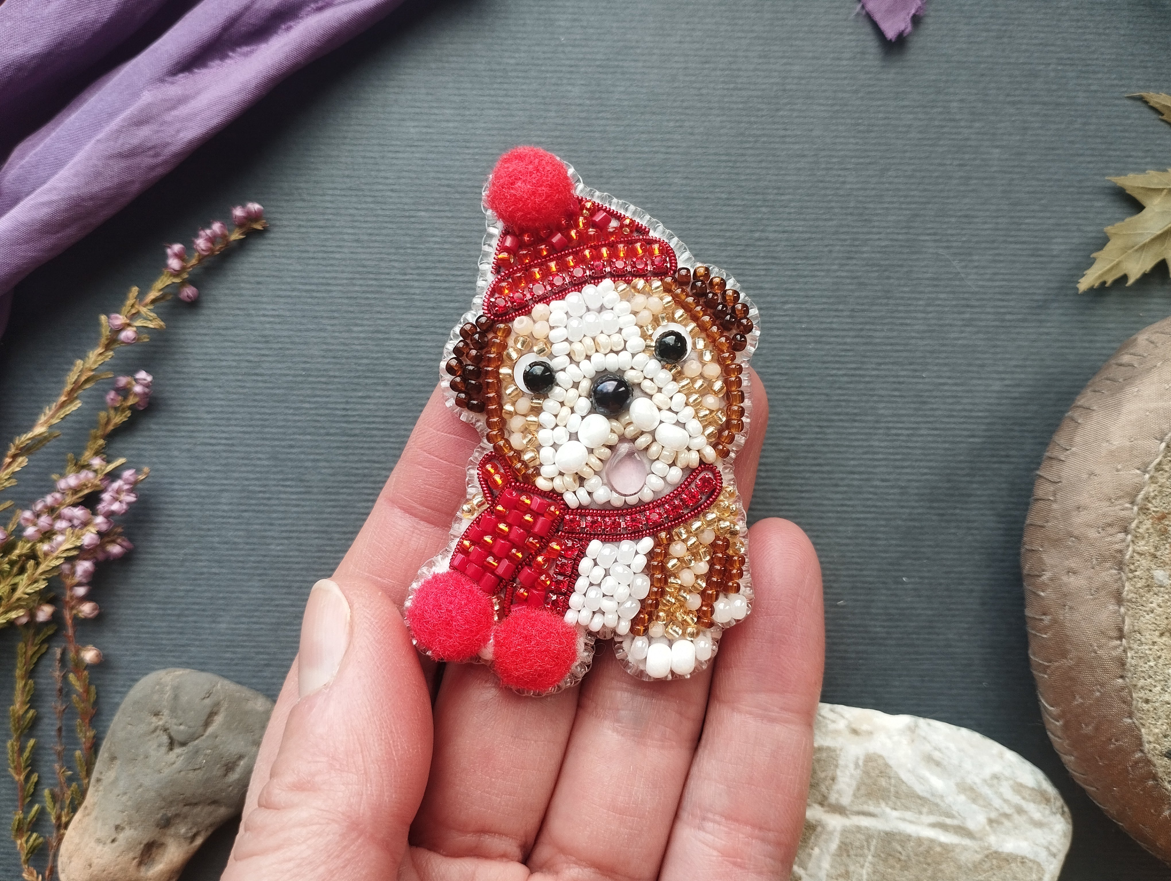 A hand holding a small, intricately beaded embroidered dog wearing a red hat and scarf.