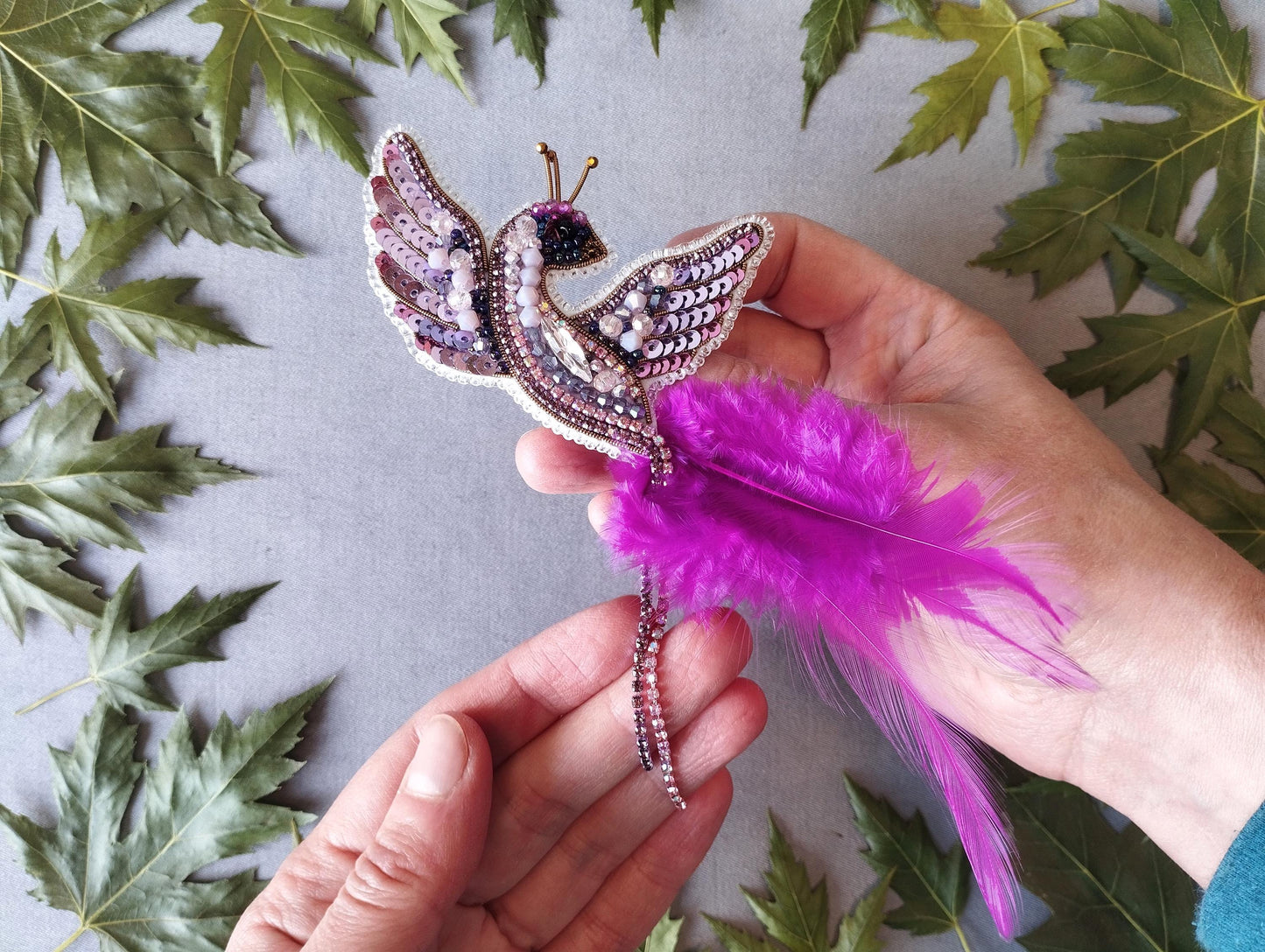 A hand holding a decorative beaded bird-shaped brooch with a feather attached, surrounded by green leaves.