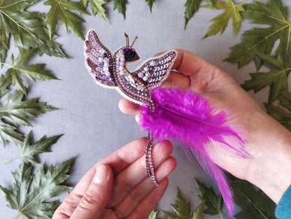 A hand holding a decorative beaded bird-shaped brooch with a feather attached, surrounded by green leaves.