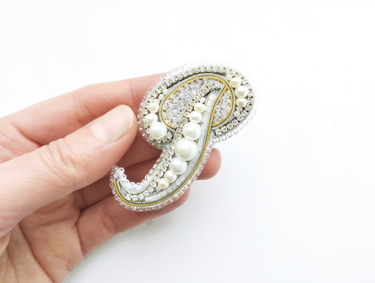 A hand holding a decorative piece of jewelry, which appears to be a brooch or pin, against a white background.