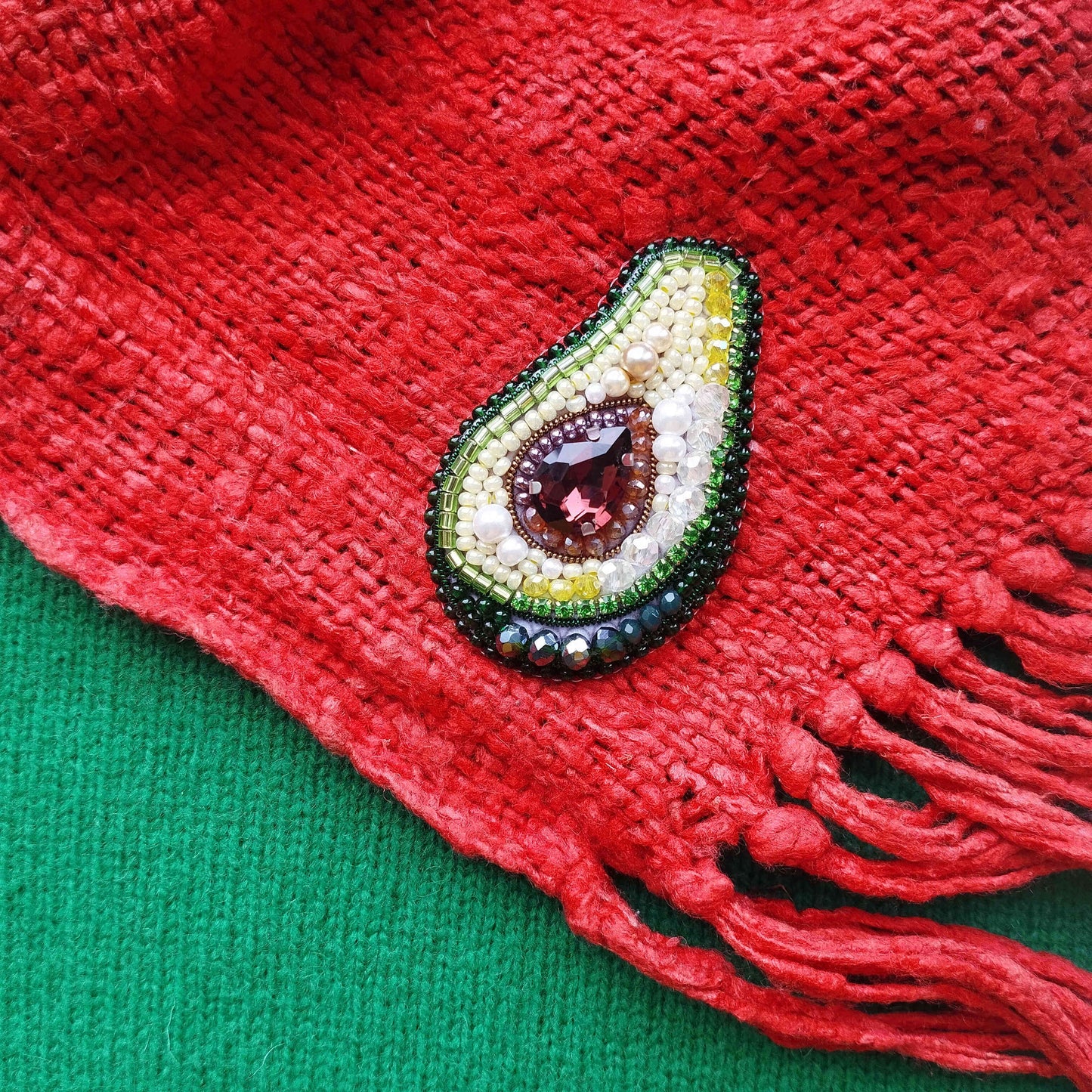 A close-up image of a red fabric with a decorative beaded avocado-shaped brooch featuring a gemstone in the center.