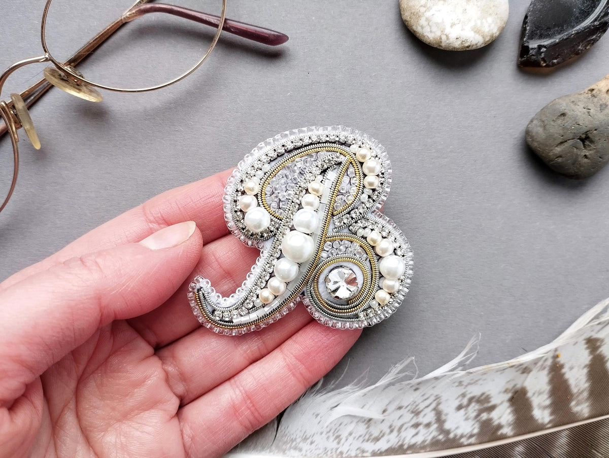 A hand holding a decorative monogram brooch with pearls and gemstones, surrounded by various objects such as glasses, rocks, and feathers.