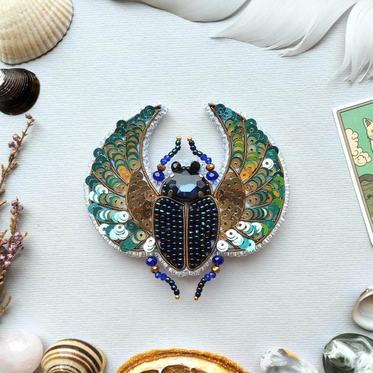a detailed and intricate embroidered piece of jewelry, resembling a large, colorful insect or beetle, surrounded by various natural elements such as seashells, feathers, and other decorative objects.