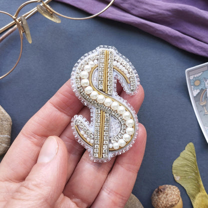 A hand holding a decorative dollar sign pin made of beads and pearls against a blue background.