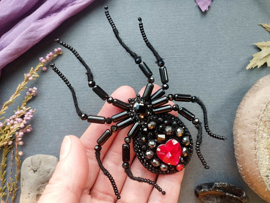 A hand is holding a black spider-shaped brooch adorned with red gemstones and black beads, set against a gray background with various natural elements such as leaves and flowers.