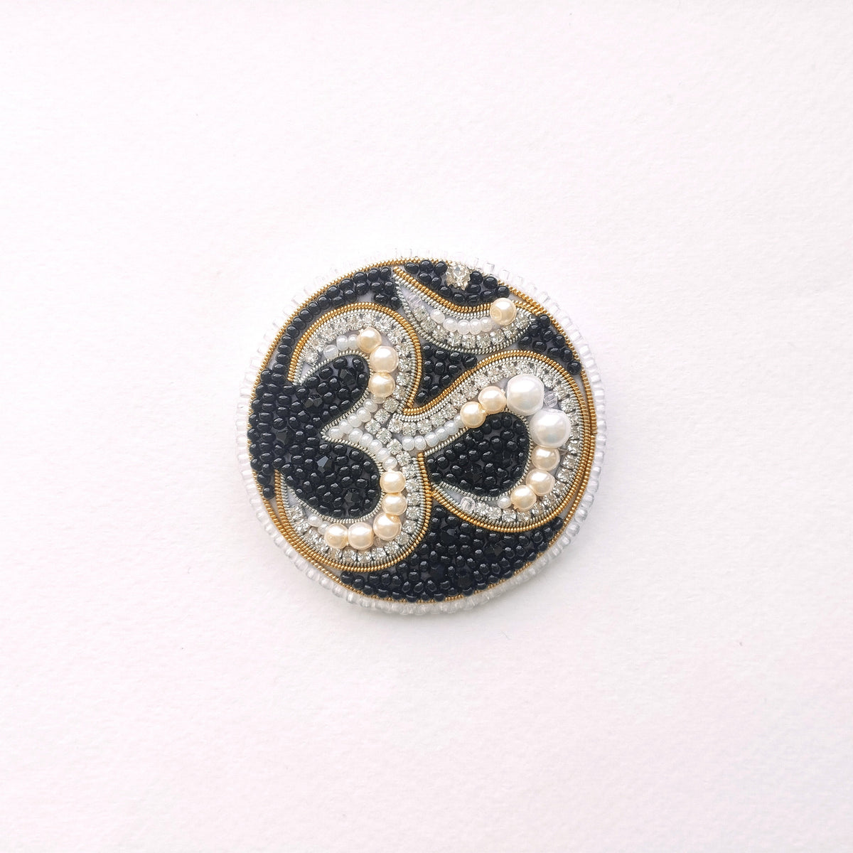 A circular, ornate piece of jewelry with a black and gold design, featuring the Om symbol and pearls.