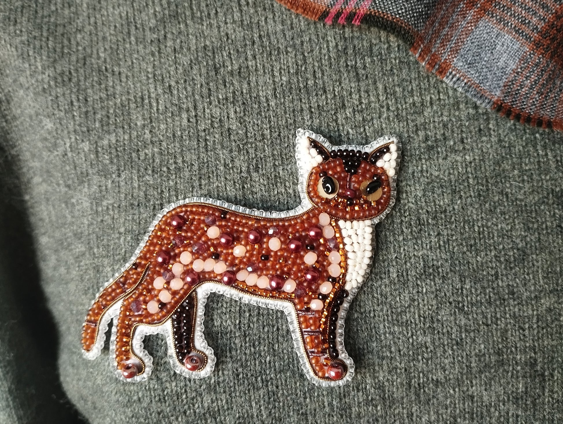 A close-up image of a brown and white embroidered cat-shaped brooch or pin, which is attached to a grey sweater.