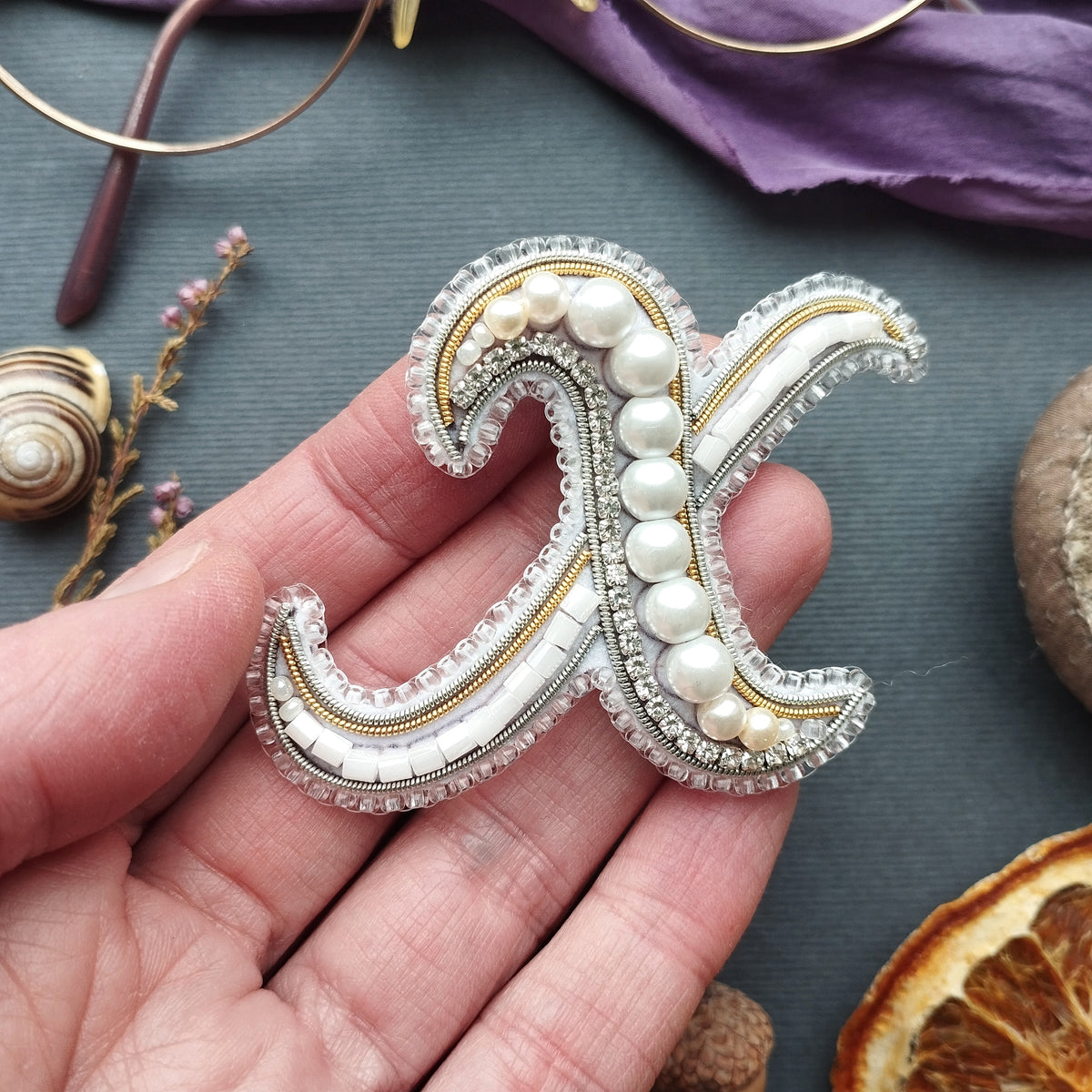 A hand holding a decorative letter 'X' made of beads and pearls against a dark background.