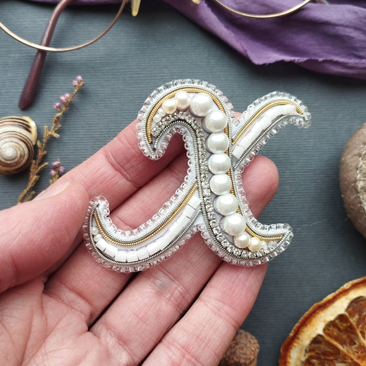 A hand holding a decorative letter 'X' made of beads and pearls against a dark background.