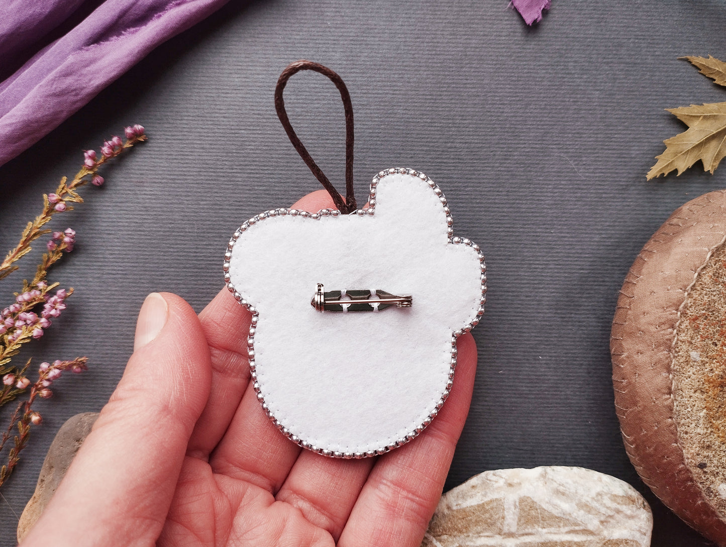 A hand holding a white felt object with a silver pin attached to it, surrounded by various natural elements such as leaves, flowers, and a mushroom.