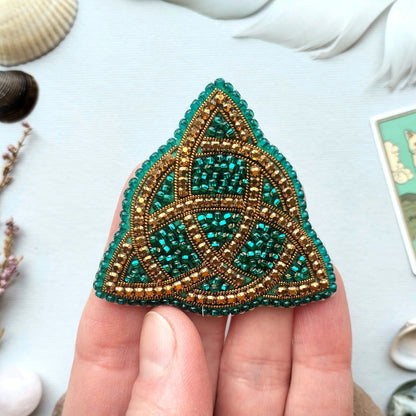 A hand holding a triangular-shaped, intricately beaded object with a green and gold color scheme.