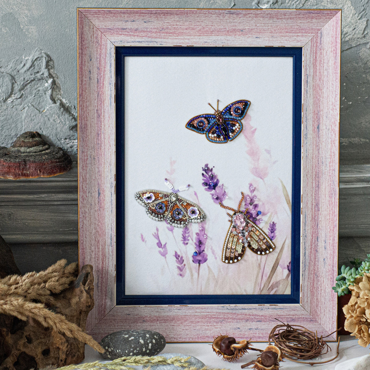 a framed artwork depicting three colorful butterflies, with one butterfly in the center and two others fluttering around it. The butterflies are adorned with intricate beading and the background is a floral pattern. The artwork is displayed on a wooden shelf, surrounded by various natural elements such as rocks, plants, and a mushroom.