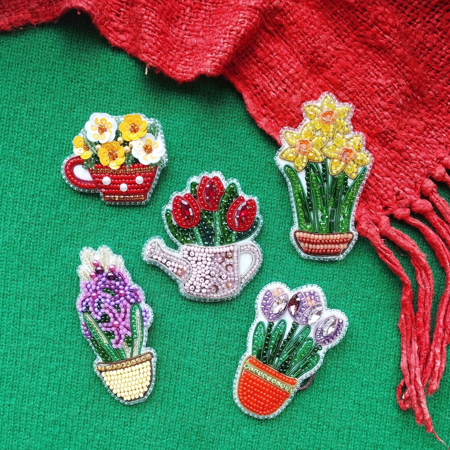 five colorful beaded brooches or pins, each depicting a different type of flower or plant, arranged on a green background.