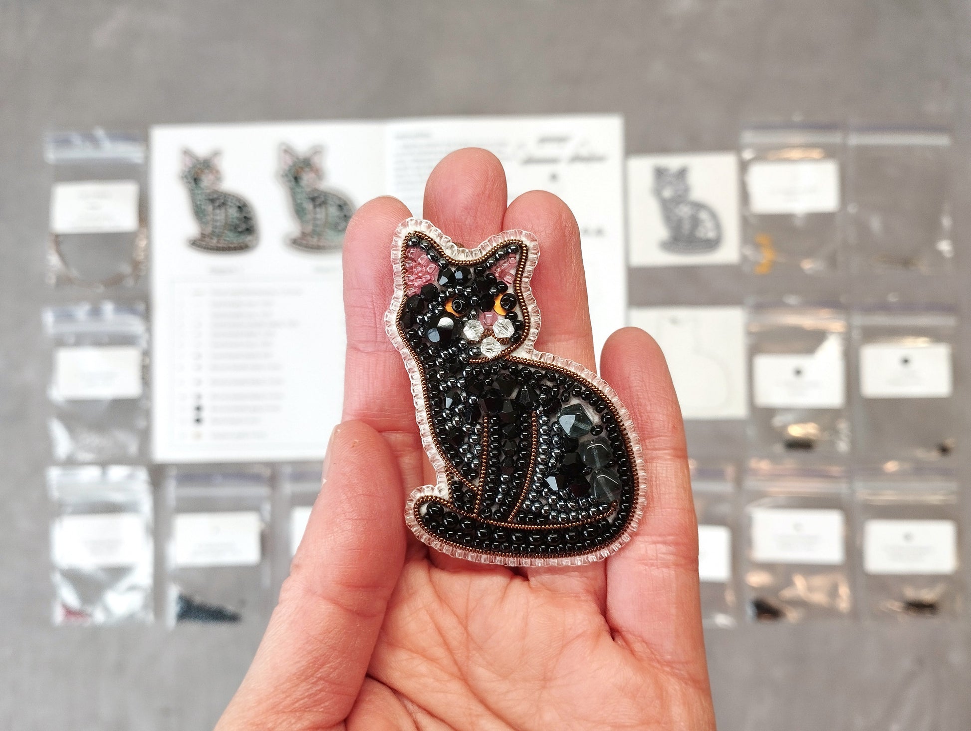 A hand holding a black cat-shaped embroidered brooch against a background of various small items and a printed page.