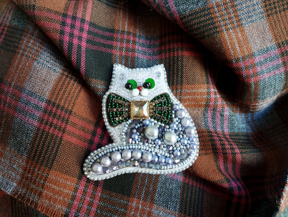 A decorative cat brooch with a green bow tie and beaded embellishments, resting on a plaid fabric background.