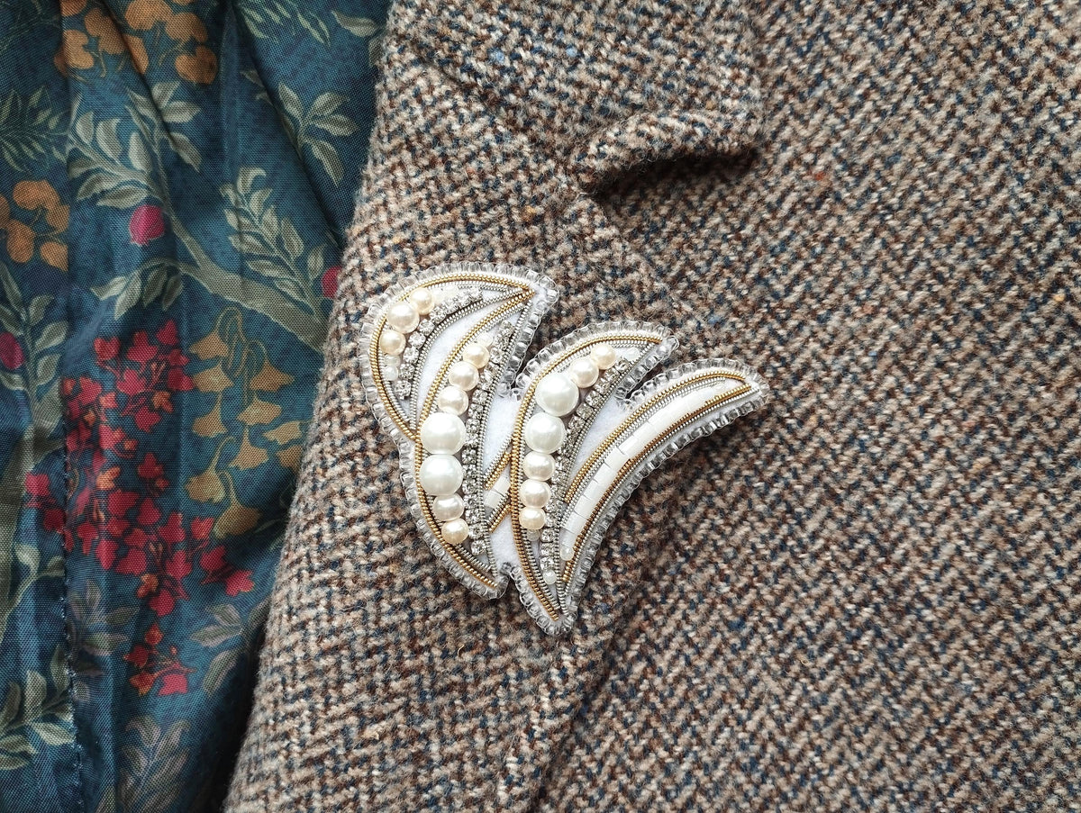 a close-up view of a decorative brooch or pin with pearls and a floral design, placed on a tweed jacket or fabric with a floral pattern.