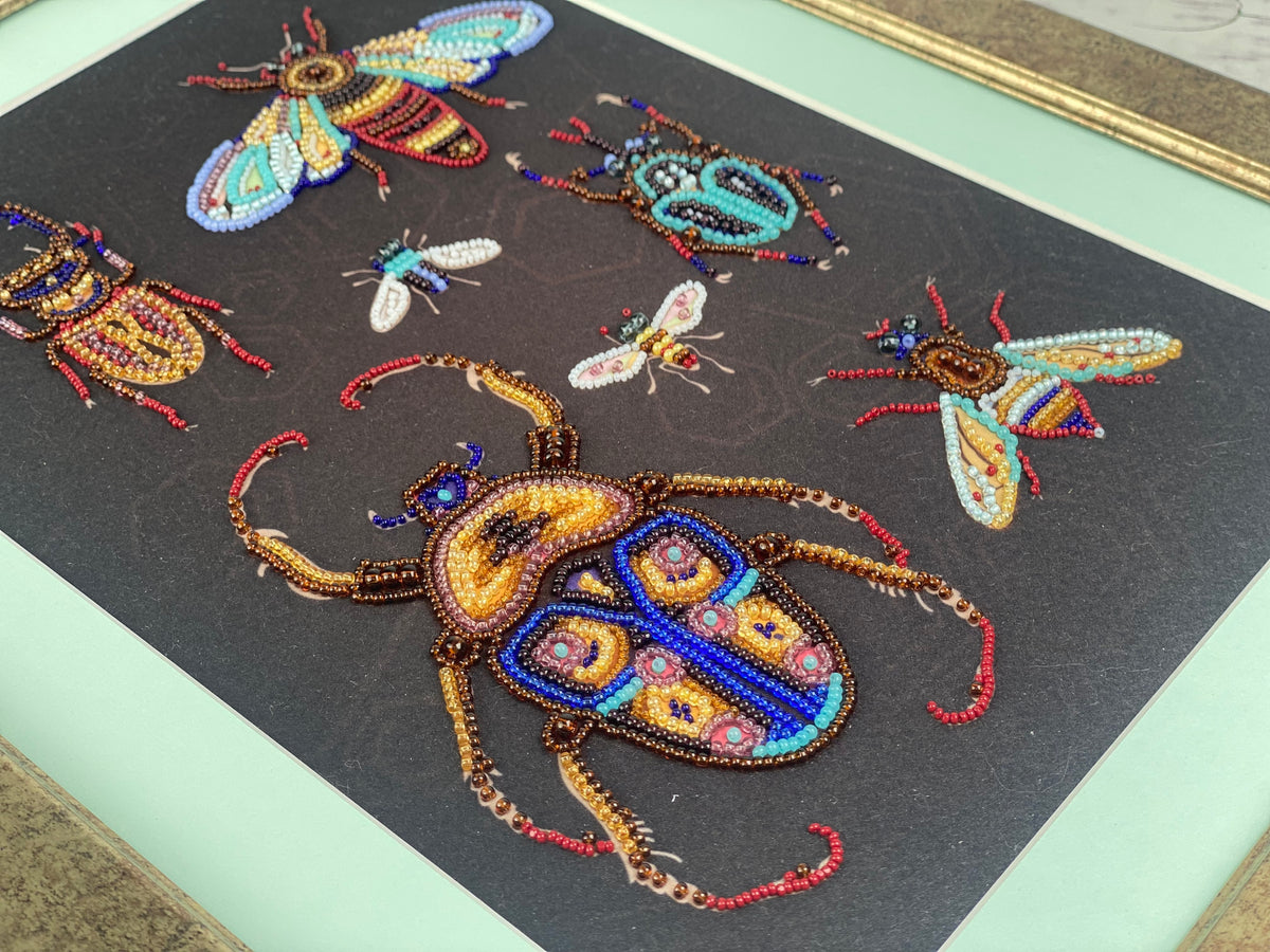 The image is a framed artwork featuring a detailed and colorful beaded design of various insects, including a large beetle with a blue and yellow pattern.