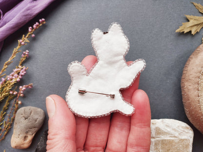 A hand holding a white fabric pin or badge with a silver pin attached to it, surrounded by various natural elements such as leaves, flowers, and rocks.