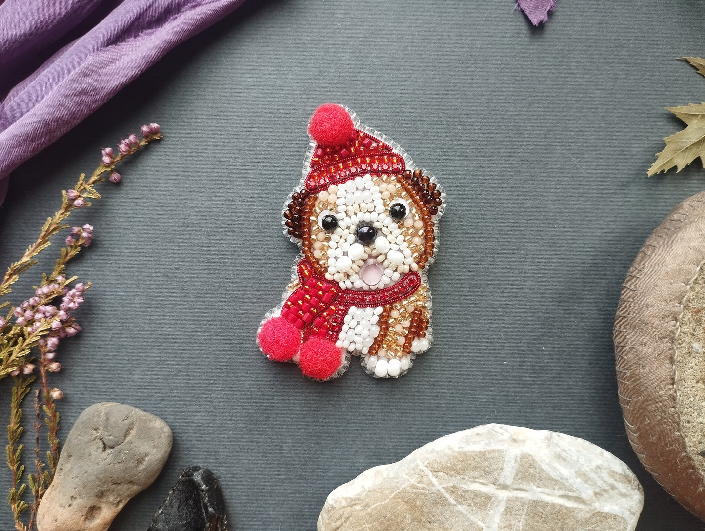 a small, intricately designed embroidered dog pin or brooch, surrounded by various objects such as rocks, leaves, and flowers, creating a visually appealing and diverse composition.
