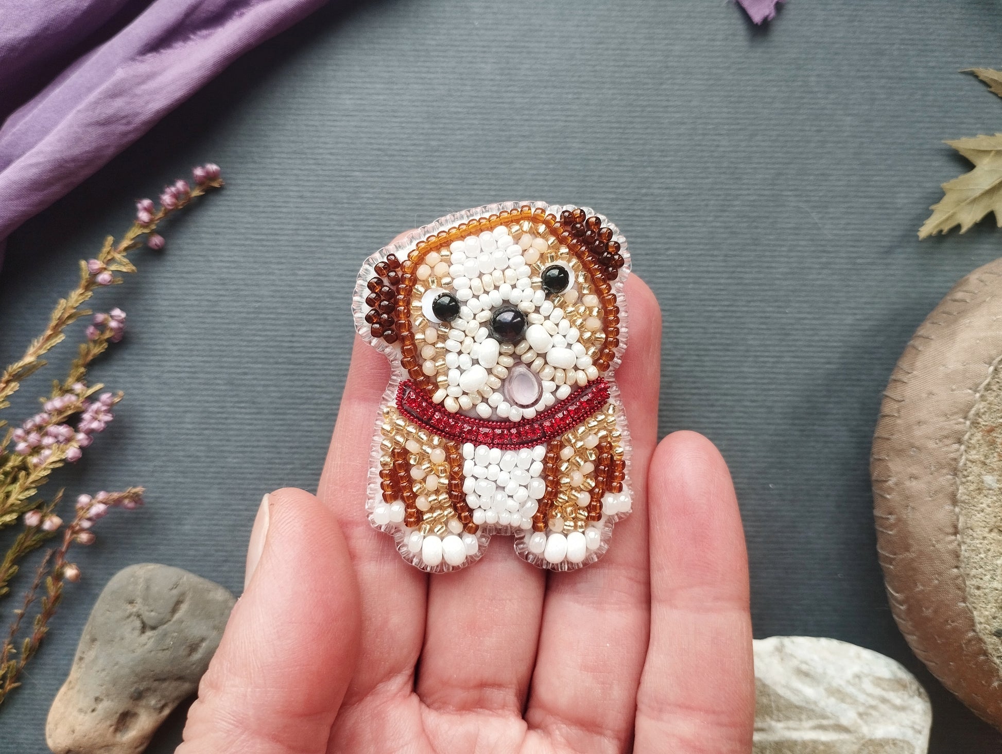 A hand holding a small, intricately beaded dog-shaped brooch against a gray background.