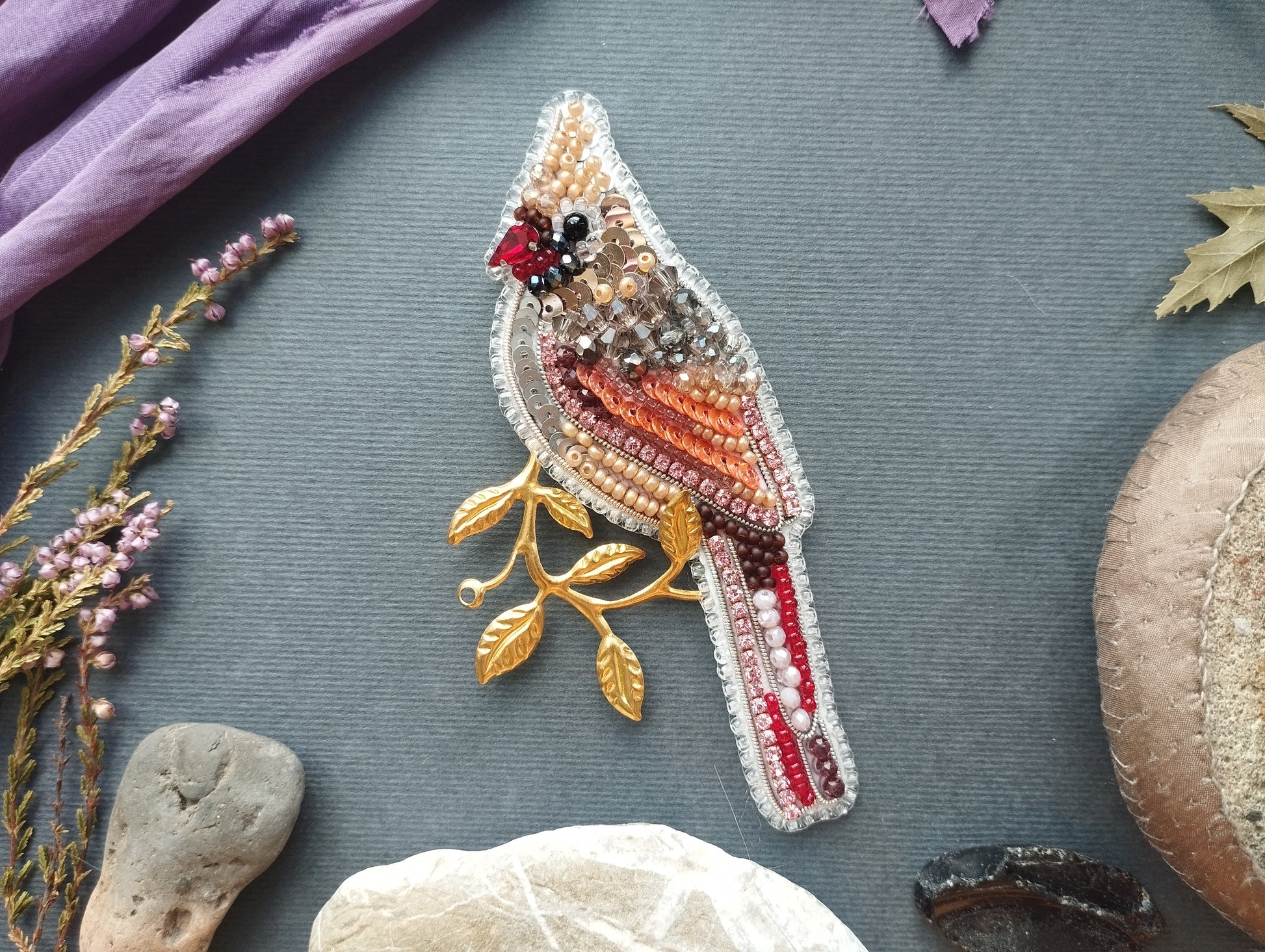 a decorative bird brooch with a beaded design, placed on a gray background among various natural elements such as leaves, rocks, and flowers.