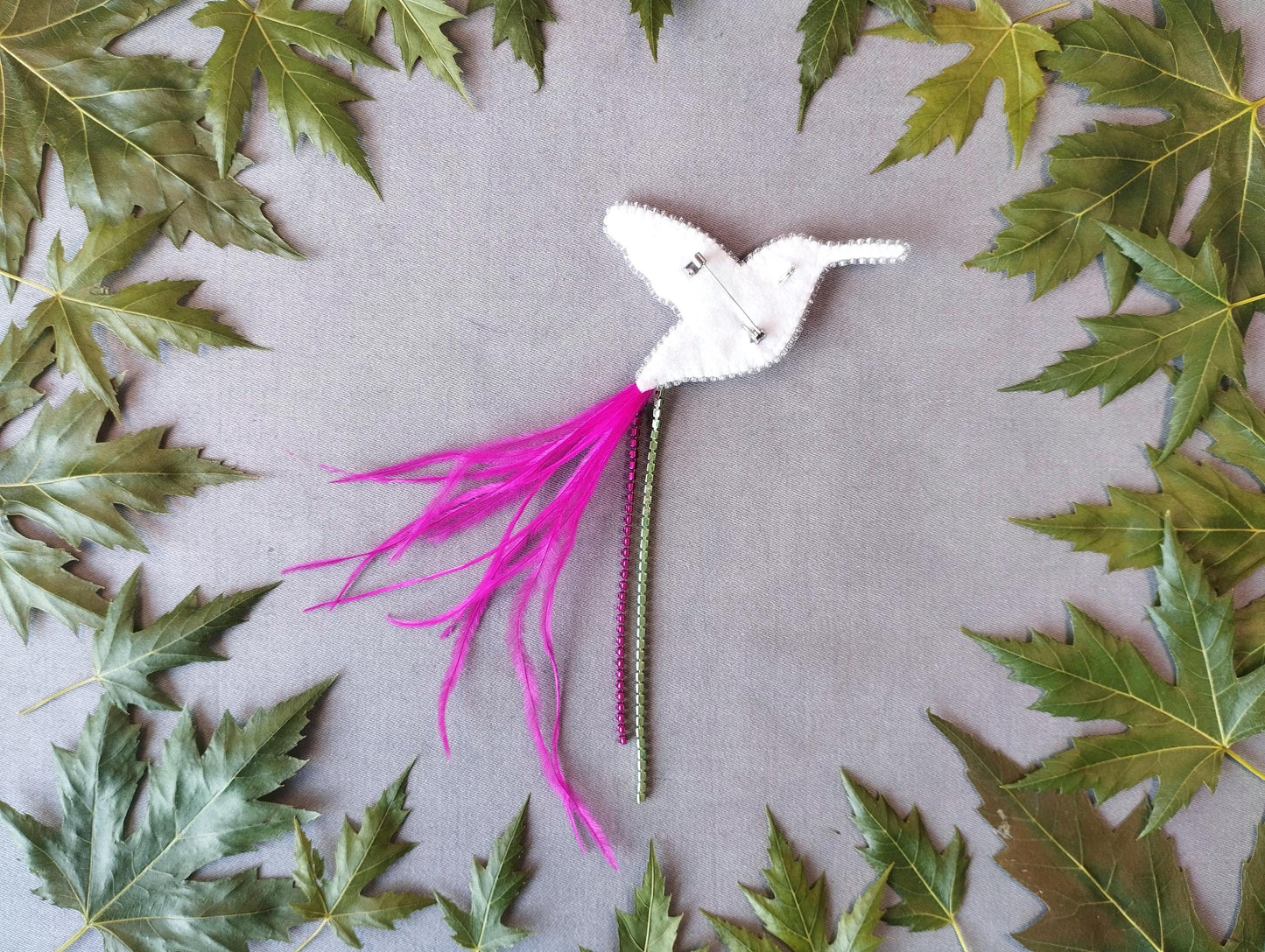 A white bird-shaped object with pink feathers and a green stem is surrounded by green leaves on a gray background.