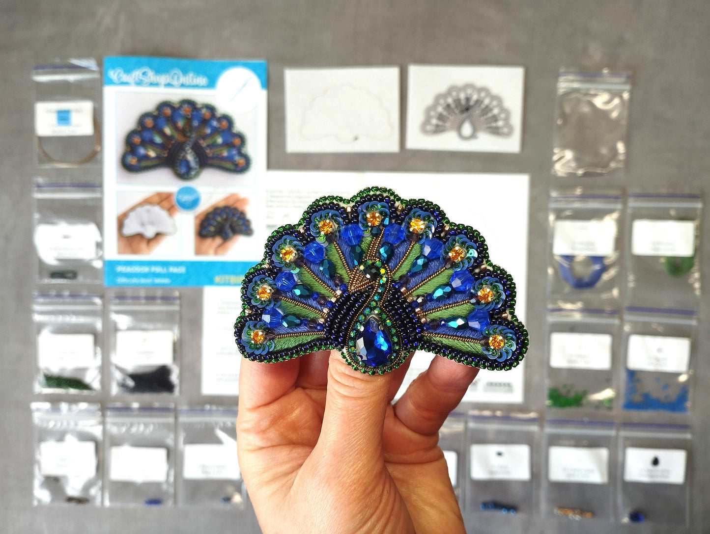 A hand holding a decorative peacock feather-shaped object, with various craft supplies and materials visible in the background.