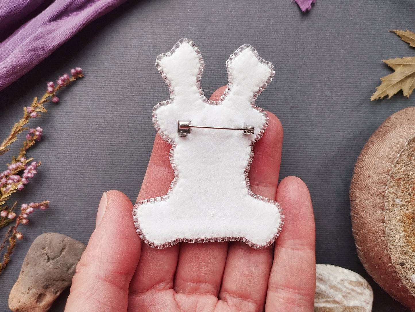 A hand holding a white felt reindeer pin against a gray background with various natural elements, such as leaves and rocks.