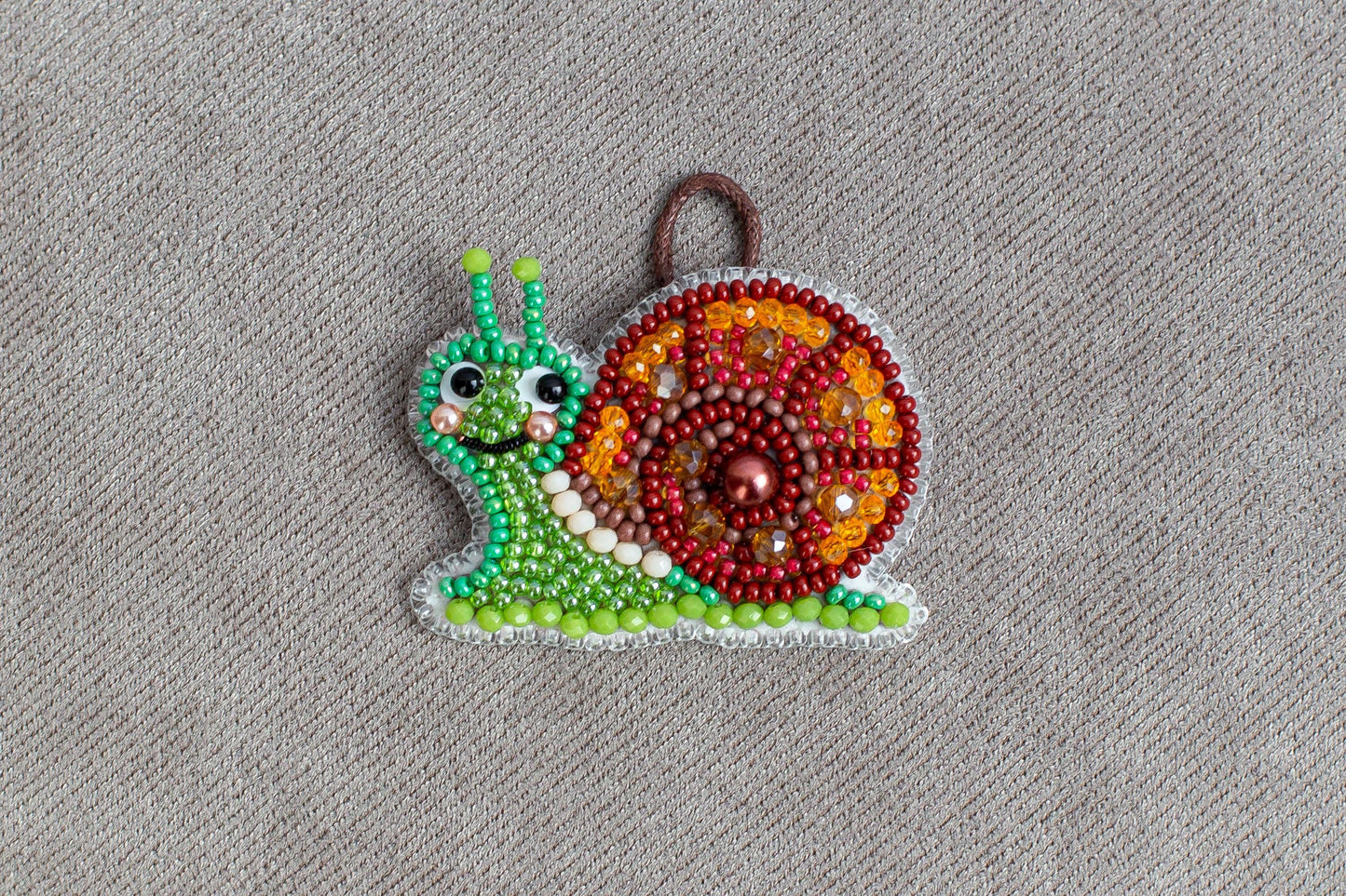 A colorful, handcrafted ornament in the shape of a snail made of beads and other materials, displayed on a textured gray background.