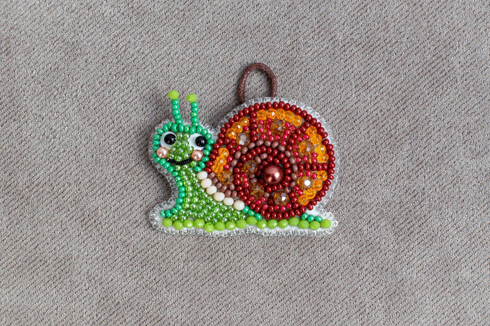 A colorful, handcrafted ornament in the shape of a snail made of beads and other materials, displayed on a textured gray background.