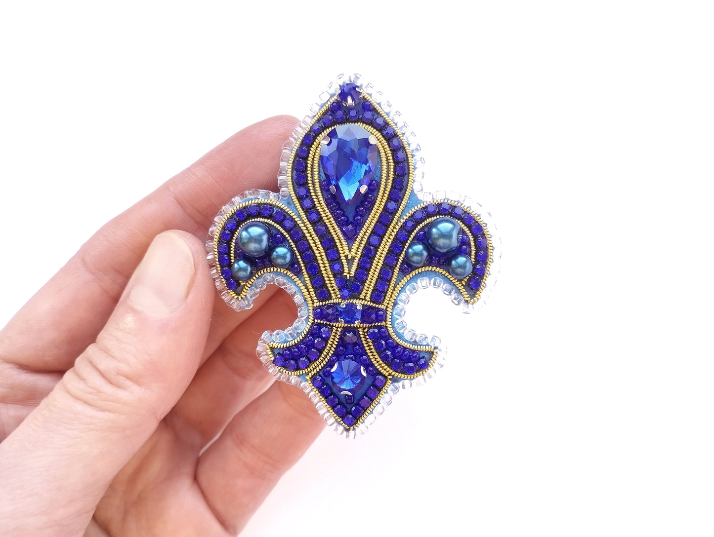 A hand holding a decorative blue and gold brooch with a fleur-de-lis design and sparkling gemstones.